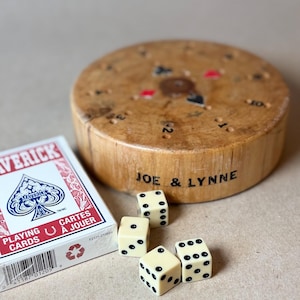 May include: A round wooden game board with a central spinner, the names JOE & LYNNE printed on the side, and card suit symbols. Next to it is a white box of Maverick playing cards and four cream-colored dice with black pips on the surface.