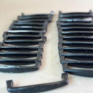 May include: A collection of black drawer pulls arranged in two parallel rows, with one additional pull placed horizontally in the foreground. Each pull features a curved handle design with two mounting points on the ends for attachment.