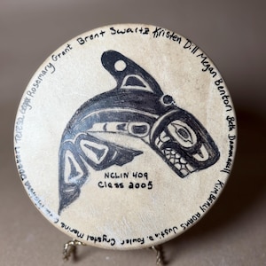 May include: A round, beige drum with a black orca design in the center. The drum has the text "NCLIN 409 Class 2005" and names around the edge. The drum is displayed on a small, gold-colored stand.