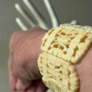 May include: A cream-colored bracelet featuring intricate, openwork carvings of repeating geometric and floral motifs. The wide, segmented band is constructed from connected panels that wrap around the wrist, showcasing detailed relief patterns.
