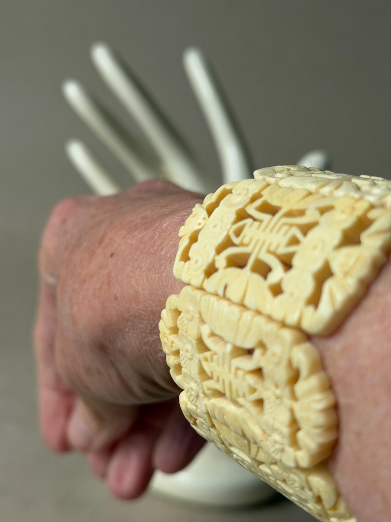 May include: A cream-colored bracelet featuring intricate, openwork carvings of repeating geometric and floral motifs. The wide, segmented band is constructed from connected panels that wrap around the wrist, showcasing detailed relief patterns.