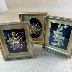 May include: Three rectangular gold-toned frames containing dried floral arrangements on black backgrounds. The left frame features a bouquet with a pink ribbon, the center frame has an oval mat with a spiky plant, and the right frame holds a curved arrangement.