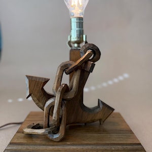 May include: A wooden anchor lamp with a decorative Edison-style bulb. The anchor and chain are carved from wood and sit on a rectangular wooden base. The lamp has a nautical theme and is a warm brown color.