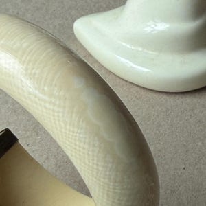 May include: A thick, off-white bangle bracelet featuring a subtle, cross-hatched, textured surface pattern. The bracelet has a smooth, rounded exterior profile and a solid, lighter-colored interior band, positioned near a white ceramic object.
