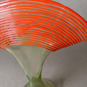 May include: A decorative glass art piece featuring a flared, trumpet-shaped base in clear and green hues, supporting a vibrant orange, layered, circular design. The piece has a modern, artistic aesthetic.