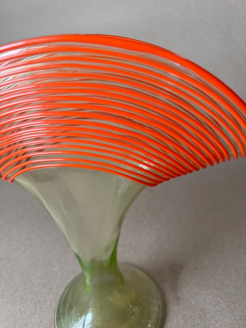 May include: A decorative glass art piece featuring a flared, trumpet-shaped base in clear and green hues, supporting a vibrant orange, layered, circular design. The piece has a modern, artistic aesthetic.