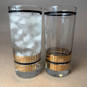 May include: Two tall, clear drinking glasses with black bands at the top and gold vertical stripes near the base. One glass is filled with ice and water. The glasses are on a neutral background.