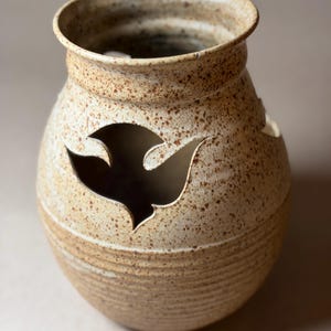 May include: A speckled, beige ceramic vase with a wide opening and a rounded body. The vase features a cut-out design, resembling a bird in flight. The exterior has a textured, rustic appearance, with horizontal ridges near the base.