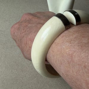 May include: A thick, off-white, curved bangle bracelet featuring a smooth, polished finish. The bracelet is designed with a wide, open gap at the top, which is accented by two dark, metallic-toned end caps that provide a contrast to the light material.