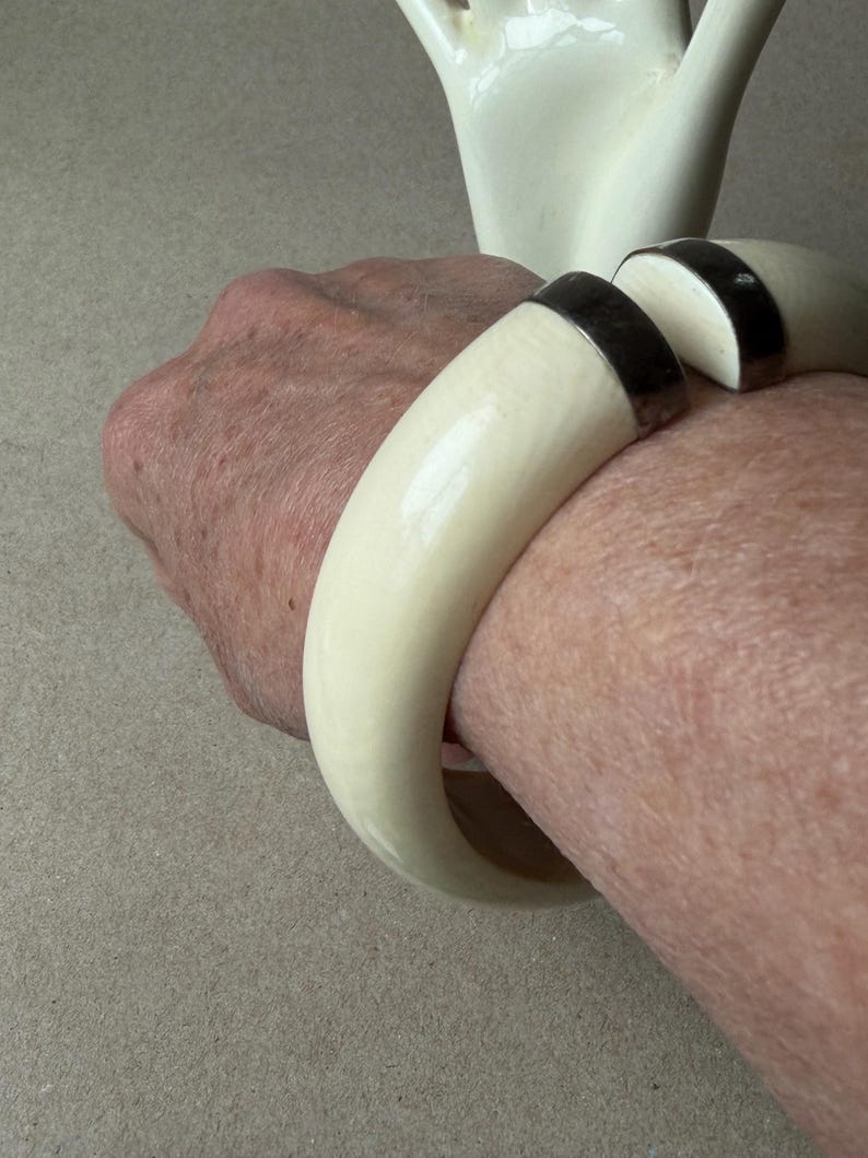 May include: A thick, off-white, curved bangle bracelet featuring a smooth, polished finish. The bracelet is designed with a wide, open gap at the top, which is accented by two dark, metallic-toned end caps that provide a contrast to the light material.