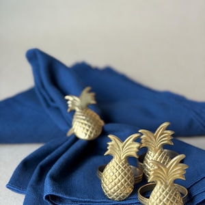 May include: Gold-colored pineapple napkin rings on a folded blue napkin. The pineapple rings have a textured surface and are arranged in a decorative display. The napkin is a deep shade of blue.