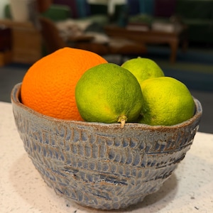 May include: A ceramic bowl with a textured blue exterior holds an orange and three green limes. The bowl has a rustic, handcrafted appearance. The fruit is fresh and vibrant, suggesting a healthy snack or decorative display.