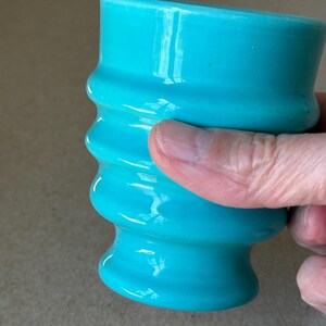 May include: A bright turquoise ceramic tumbler featuring a series of horizontal, raised ridges along its cylindrical body. The vessel has a smooth, glossy finish and a slightly flared base, providing a textured grip for the user.