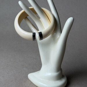 May include: A cream-colored open cuff bracelet with a smooth, rounded profile. The bracelet features two polished silver-tone metal end caps at the opening, providing a clean contrast to the light-toned band. It is displayed on a white hand form.