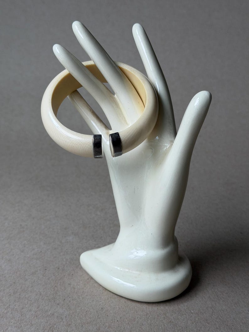 May include: A cream-colored open cuff bracelet with a smooth, rounded profile. The bracelet features two polished silver-tone metal end caps at the opening, providing a clean contrast to the light-toned band. It is displayed on a white hand form.