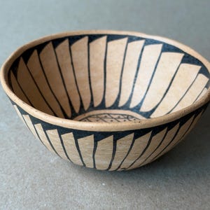 May include: A handmade ceramic bowl with a beige exterior and a black geometric pattern. The interior features a similar design. The bowl's rim is outlined in black, and the overall aesthetic is rustic and artistic. This bowl is perfect for serving food or as a decorative piece.