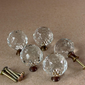 May include: Four spherical, clear, faceted glass cabinet knobs with metallic bases. Each knob features a multi-faceted surface that reflects light. Included are four matching metal mounting screws with threaded shafts and flat heads.