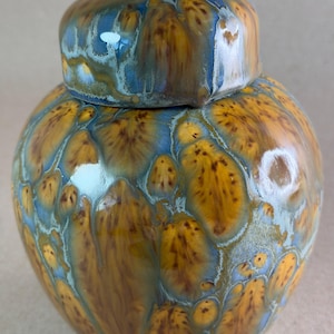 May include: A ceramic jar with a lid featuring a crystalline glaze pattern. The surface displays a blend of golden-brown, amber, and light blue colors with organic, petal-like shapes and dark brown speckles throughout the glossy, smooth finish.