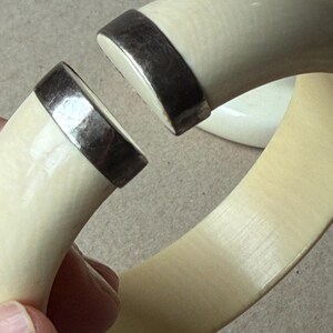 May include: A thick, off-white, curved bangle bracelet featuring a wide, open gap. The two ends of the bracelet are capped with polished, dark metallic silver-toned hardware, creating a distinct contrast against the smooth, light-colored material.