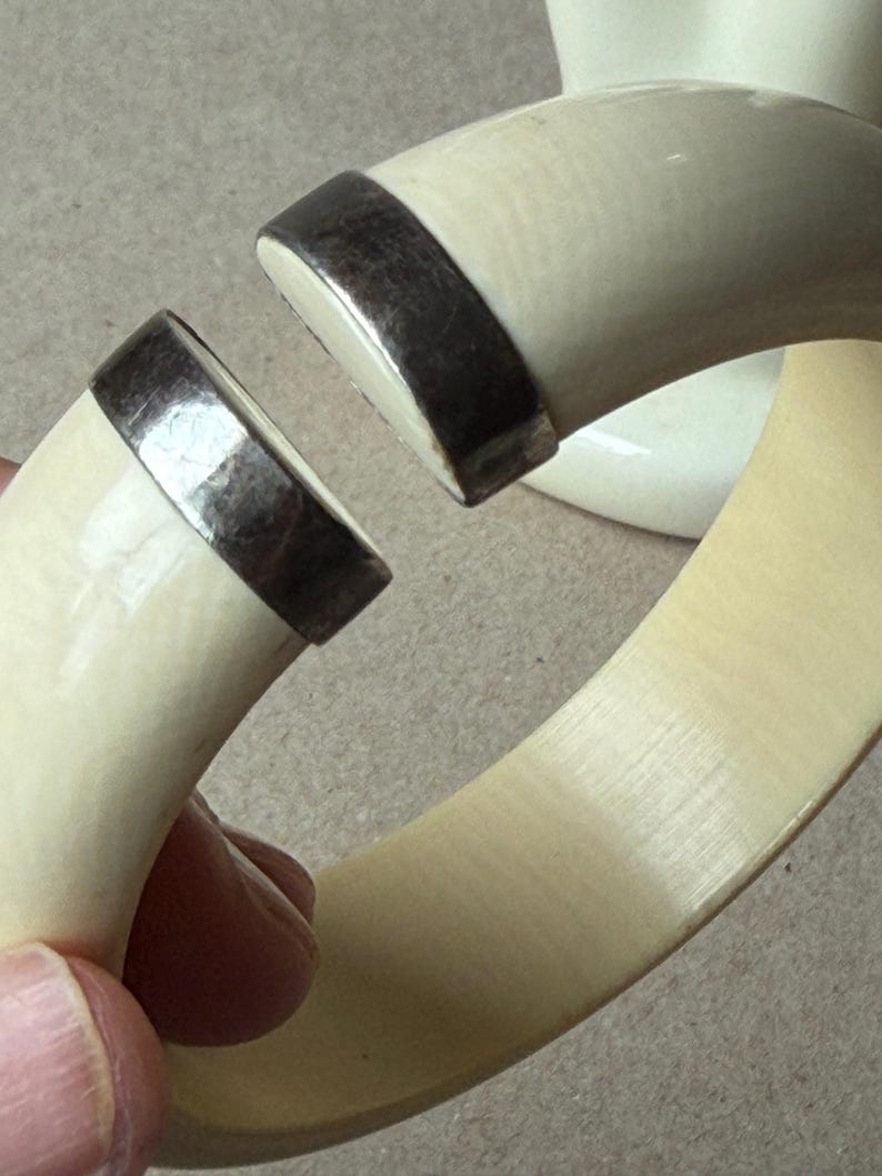 May include: A thick, off-white, curved bangle bracelet featuring a wide, open gap. The two ends of the bracelet are capped with polished, dark metallic silver-toned hardware, creating a distinct contrast against the smooth, light-colored material.