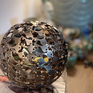 May include: A decorative sphere composed of numerous metal keys, creating a unique, textured surface. The sphere is a metallic silver color and sits on a stack of papers. The background is blurred, with hints of other decorative items.