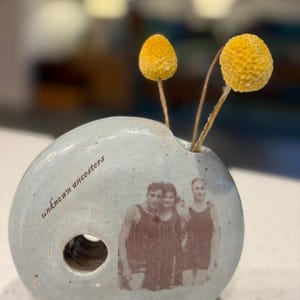 May include: A light blue ceramic vase with a circular design, featuring a black and white photo of three people in swimwear. The vase has a hole and the words "unknown ancestors" printed on it. Two yellow, spherical flowers are in the vase.