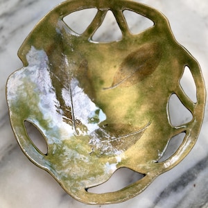 May include: A handcrafted ceramic bowl with a leaf design. The bowl features a unique, openwork rim and a glossy, olive green and gold glaze. The interior showcases leaf imprints, adding a natural, artistic touch to this decorative piece.