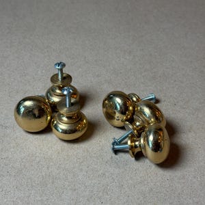 May include: A set of four round, polished brass cabinet knobs with a smooth, reflective finish. Each knob features a spherical head attached to a short, cylindrical base and includes a silver-colored metal mounting screw protruding from the back.