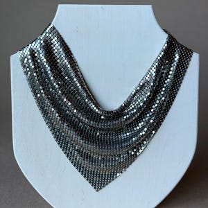 May include: A silver-colored mesh necklace displayed on a white mannequin bust. The necklace is made of small, reflective metal discs, creating a draped, V-shaped design. The necklace is a statement piece, suitable for formal occasions.