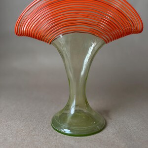 May include: A decorative glass vase with a flared top and a wide base. The top of the vase is adorned with numerous orange, curved, wire-like elements. The vase has a light green tint and a unique, artistic design.