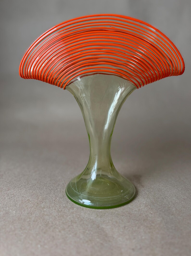 May include: A decorative glass vase with a flared top and a wide base. The top of the vase is adorned with numerous orange, curved, wire-like elements. The vase has a light green tint and a unique, artistic design.