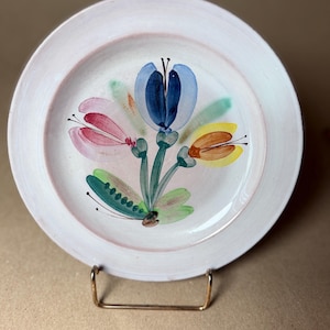 May include: A white ceramic plate with a hand-painted floral design. The flowers are in shades of blue, pink, yellow, and green. The plate has a slightly raised rim and is displayed on a gold-colored metal stand.