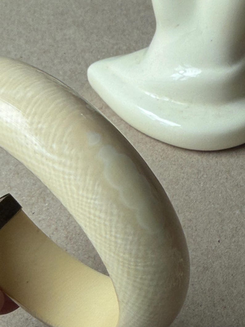 May include: A thick, curved bangle bracelet with a cream-colored, textured surface featuring a subtle cross-hatch pattern. The interior of the bracelet is a smooth, solid light yellow. A white ceramic hand-shaped display stand is visible nearby.