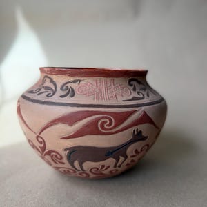 May include: A hand-painted, round, earthenware pot with a light tan base. The pot features a brown and red design of a mountain range, swirling clouds, and a stylized animal. The rim is a dark red with a decorative border.