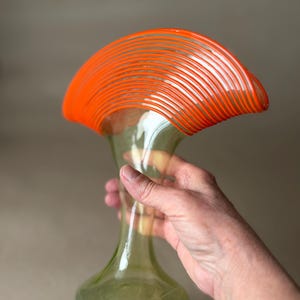 May include: A clear glass vase with a flared, orange-striped top and a slender, green-tinted body. The vase has a rounded base and is held by a hand, showcasing its elegant design. The vase is approximately 12 inches tall.