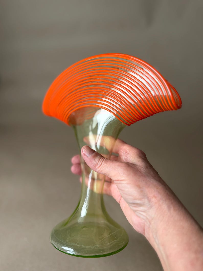 May include: A clear glass vase with a flared, orange-striped top and a slender, green-tinted body. The vase has a rounded base and is held by a hand, showcasing its elegant design. The vase is approximately 12 inches tall.