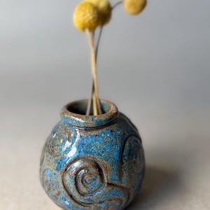 May include: A small, round, blue ceramic vase with a speckled finish and embossed spiral patterns. The vase has a short, thick neck and contains three dried, round, yellow billy ball flowers with thin, brown stems extending upwards.