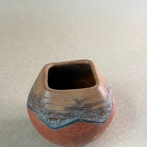 May include: A small, round, terracotta-colored clay pot with a square-shaped opening. The upper portion features a textured, dark brown band with etched geometric triangular patterns and a distinct zigzag line encircling the vessel's neck.