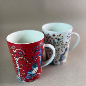 May include: Two ceramic mugs with white interiors and handles. The front mug is red with a blue owl and tree pattern. The back mug is white with a matching blue and red owl and tree pattern. Both mugs feature a tapered shape and a smooth finish.