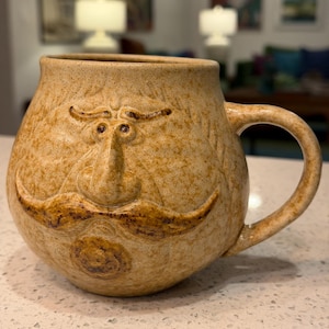 May include: A tan ceramic mug with a whimsical face design. The mug features a handle and a textured surface, with a sculpted nose, mustache, and eyes. The mug is designed to resemble a face.