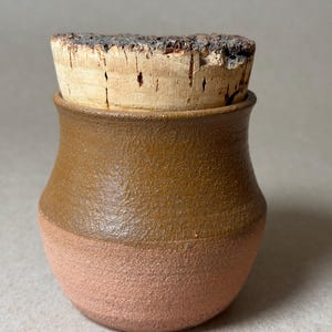 May include: A small, handcrafted ceramic jar with a cork stopper. The jar has a two-tone design, with a textured, light brown upper section and a textured, reddish-brown lower section. The cork stopper is a light brown color.