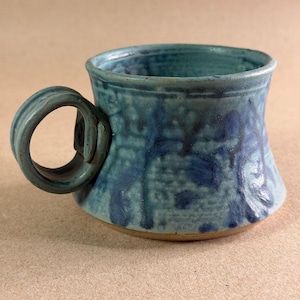 May include: A ceramic mug with a wide, flared base and a circular handle. The exterior features a mottled blue glaze with darker blue abstract patterns and a textured surface. The base of the mug shows a natural, unglazed brown clay rim.