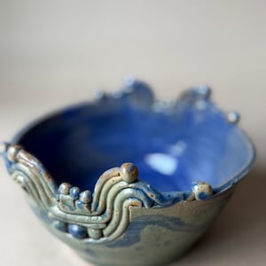 May include: A handmade ceramic bowl with a blue interior and a textured, wave-like rim. The bowl's exterior has a mottled blue-green glaze. Decorative elements include small, raised spheres and curved lines along the rim. The bowl is a unique piece of pottery.