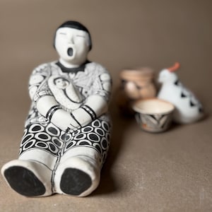 May include: A handcrafted ceramic figurine of a seated person with a baby, featuring black and white patterns. The figurine has an open mouth and is surrounded by small pottery pieces, including a bird-shaped item with an orange beak.