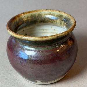 May include: A small, round ceramic pot with a wide, flared rim. The exterior features a deep, glossy burgundy and dark brown glaze, while the interior and the rim are finished in a lighter, creamy beige with mottled brown and green accents.