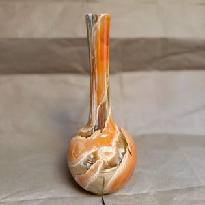 May include: A tall, slender vase with a rounded base and a narrow neck. The surface features a marbled pattern with swirling colors of bright orange, deep brown, and creamy white, creating a fluid, organic appearance across the entire exterior.