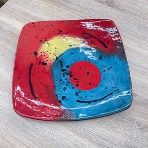 May include: Square ceramic plate with a vibrant abstract design. The plate features a red background with a yellow and blue spiral in the center. Black paint splatters and curved lines add to the artistic flair. A red circle is in the center of the blue spiral.