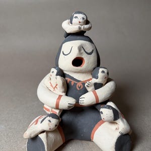 May include: A handmade ceramic sculpture of a seated figure with multiple smaller figures. The main figure is painted in black and white with red accents. The sculpture has an open mouth and closed eyes, with smaller figures on the head, arms, and lap.