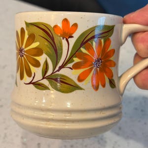 May include: Cream-colored ceramic mug with a floral design. The mug features orange and yellow flowers with green leaves. The handle is curved and the mug has a speckled finish.