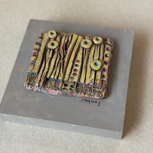 May include: A handcrafted, abstract sculpture featuring a textured, gold-colored design with vertical lines and circular accents with teal centers. The sculpture is mounted on a rectangular, gray base. The artist's signature is visible on the base.